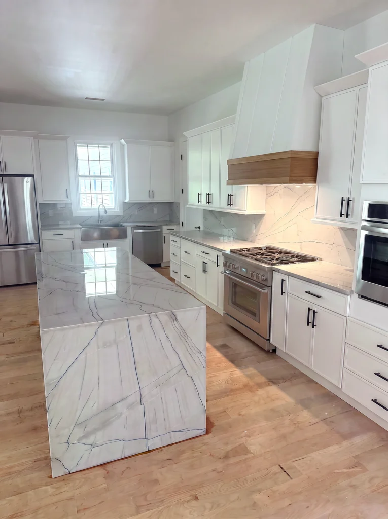 GA Granite install: quartzite waterfall island with gray-blue veins, full-height slab backsplash, white range hood with oak band and a stainless range in a white shaker kitchen